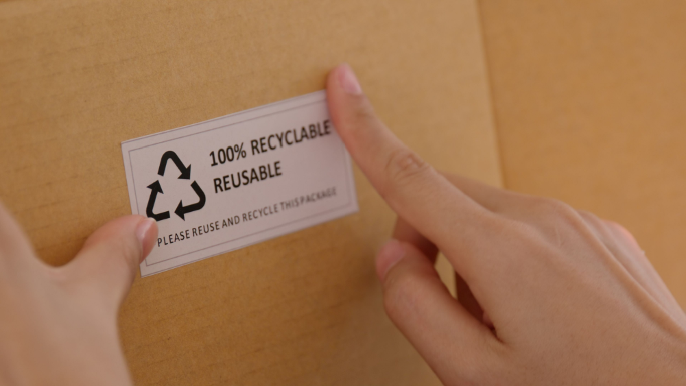 Close-up of hands applying a "100% Recyclable Reusable" label to a cardboard package.