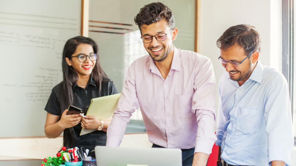 Three young Indian business people smiling and discussing something in front of a computer